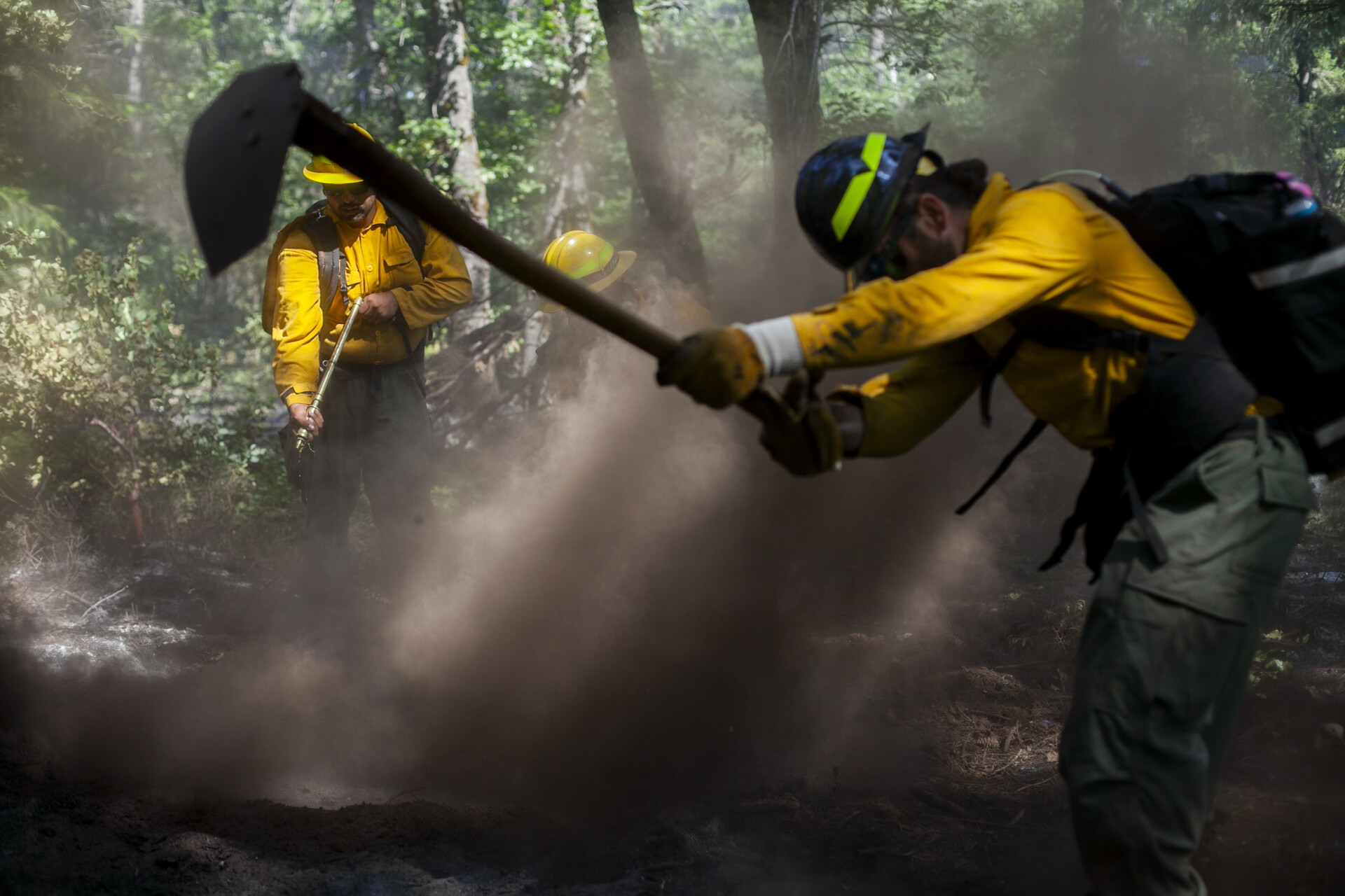 A person in a yellow jacket brings a hand tool down in a sweeping motion to the ground. 