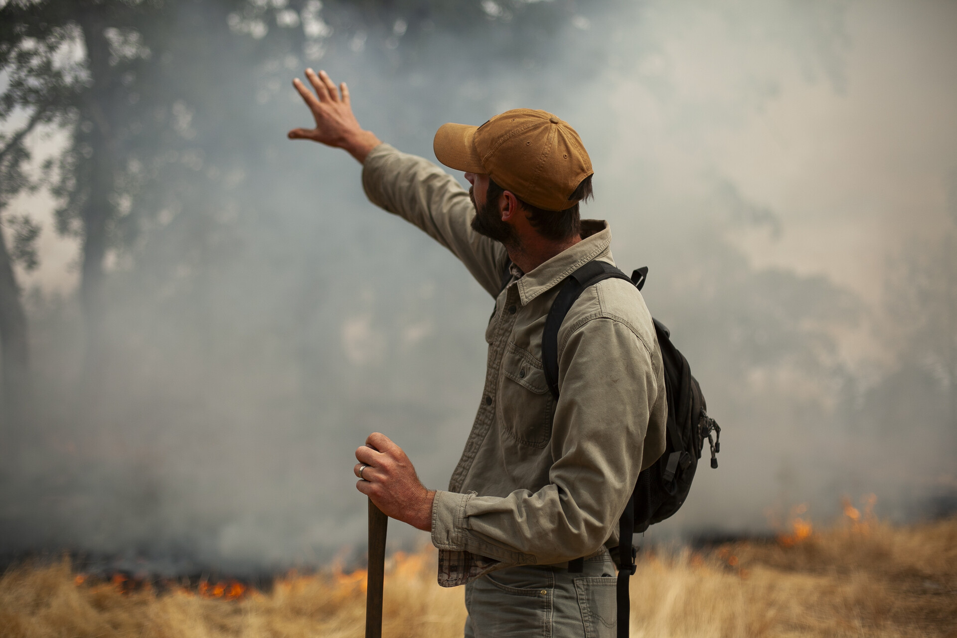A man looks up and away from the camera while holding his arm out while brush burns nearby.