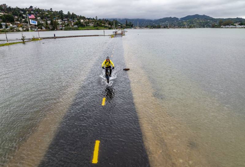 A man wearing a white bike helmet, yellow jacket and black pants rides through several inches of water on a submerged bike trail. Hills and clouds are in the foreground.