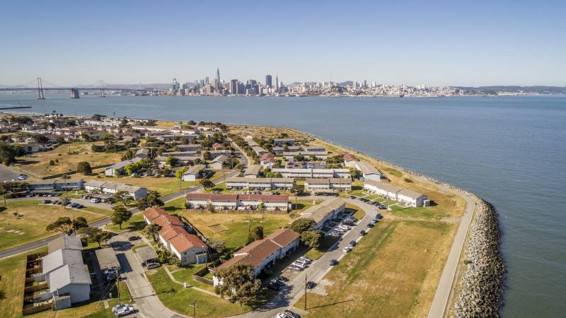 An island lined with tan apartments bordered by green grass. Water in the middleg ground and the skyline of San Francisco in the background.