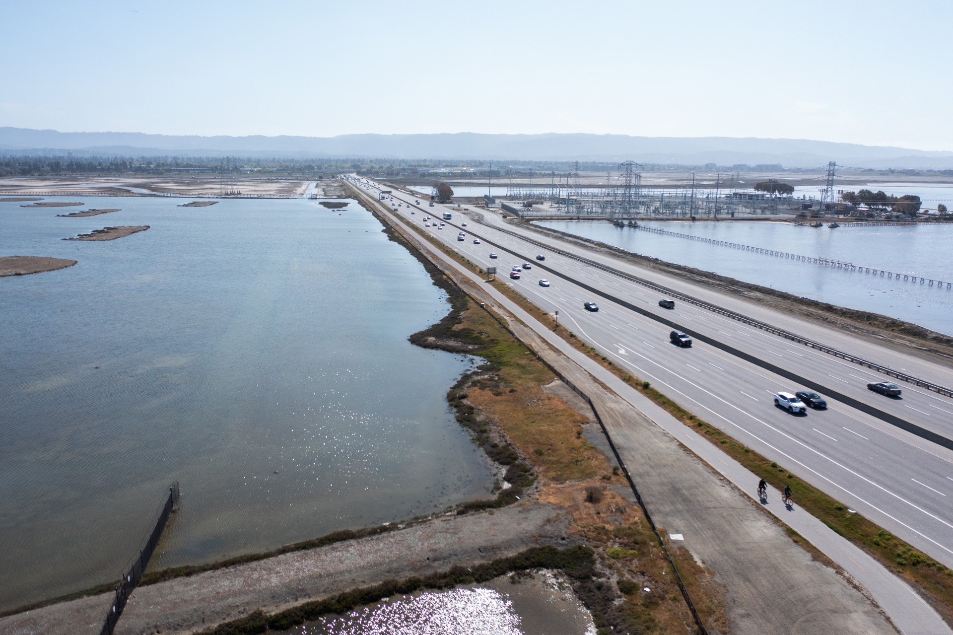 A four-lane highway with water on either side and hazy blue sky above and mirroring in the plane of water. 