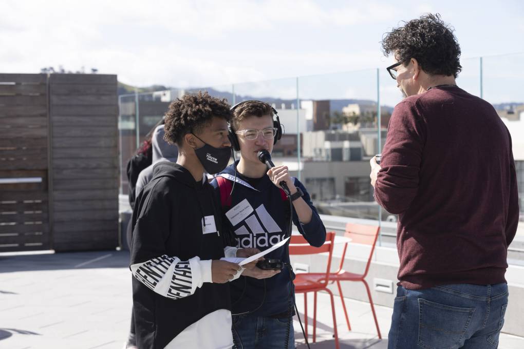 Two male students with a microphone talk with a teacher on the roof at KQED
