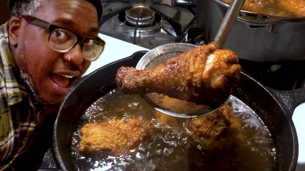A big skillet of frying chicken on the stove with a man's face next to it, looking at the camera