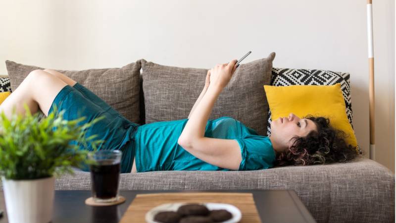 Side view shot of a young woman using digital tablet while lying down on a sofa at home with green plant, beverage and cookies on the table in front of her