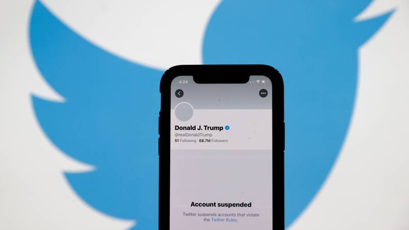 Phone screen with Donald Trump's suspended twitter account stands in front of Twitter backdrop.