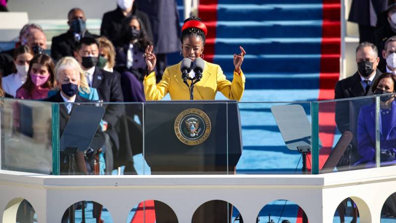 Young black woman wearing a yellow coat and red hat stands at podium with arms raised.