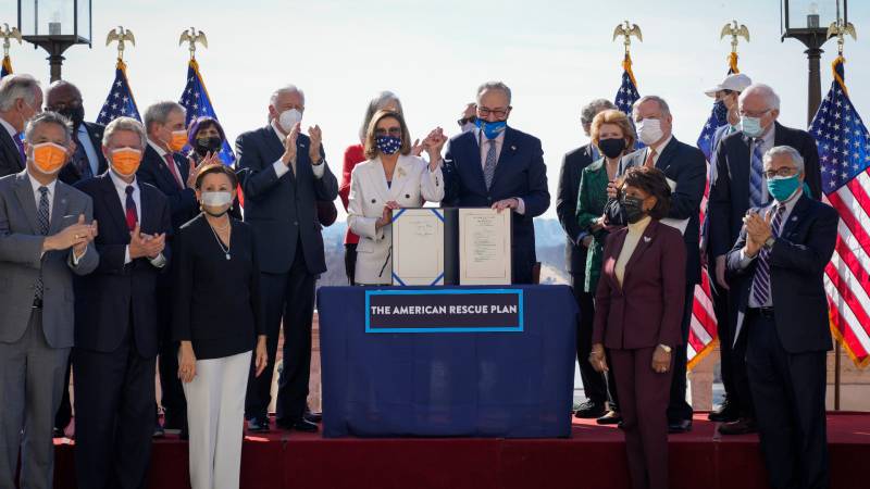Nancy Pelosi and Chuck Schumer stand behind a podium holding the covid relief bill, surrounded by members of congress