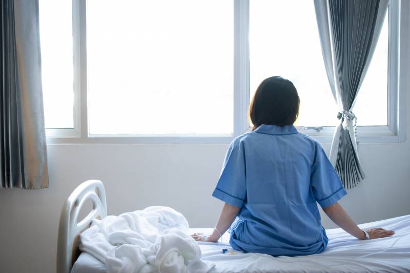 Back view of patient woman sitting on bed in hospital ward, looking away at window and hope everything will be better.