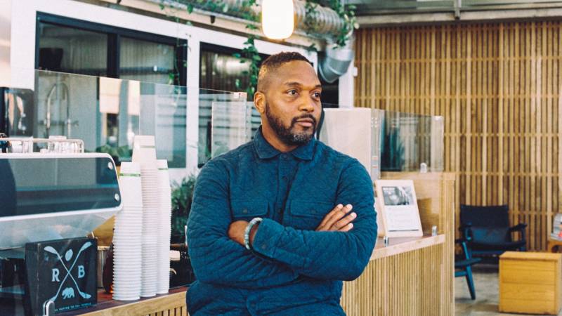A Black man is standing in front of a coffee bar at a cafe with his arms crossed. He is wearing a long sleeve, blue, button up shirt and has facial hair.