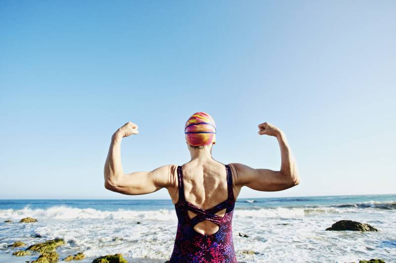 Older woman facing away from the camera flexing her biceps. She's wearing a one-piece swimsuit and a swimming cap.