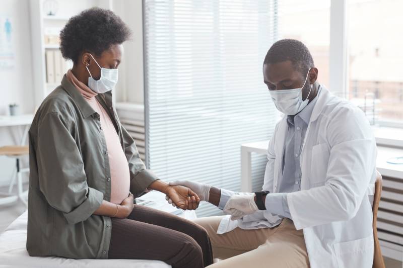 Black young male doctor in mask measuring the pulse of pregnant woman and caring about her health at hospital