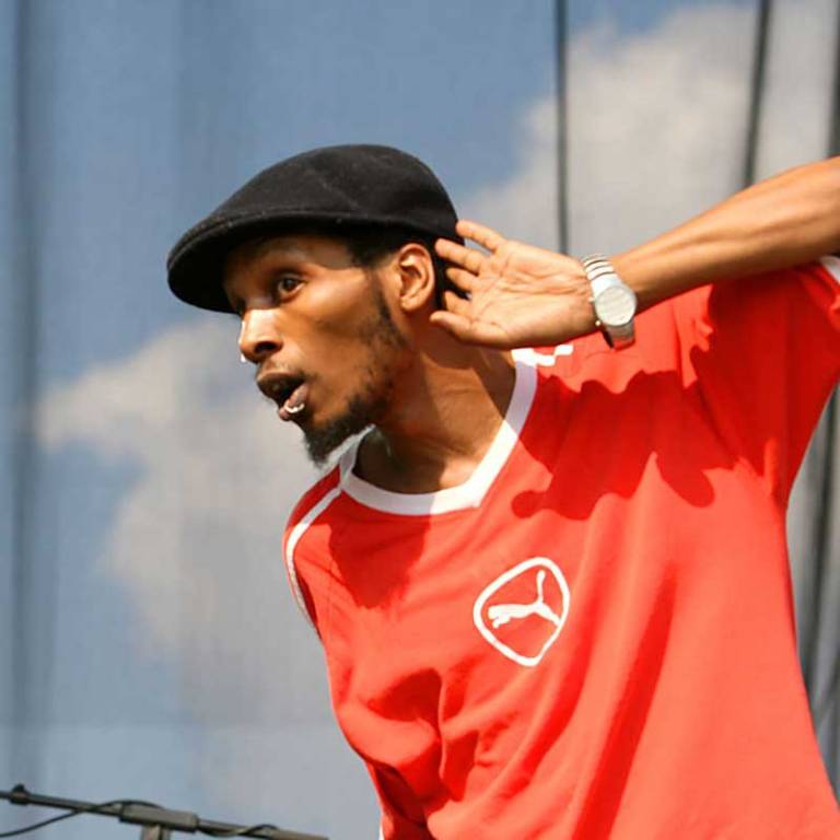 A rapper holding his hand to his ear, on stage, wearing a Kangol hat and a red shirt