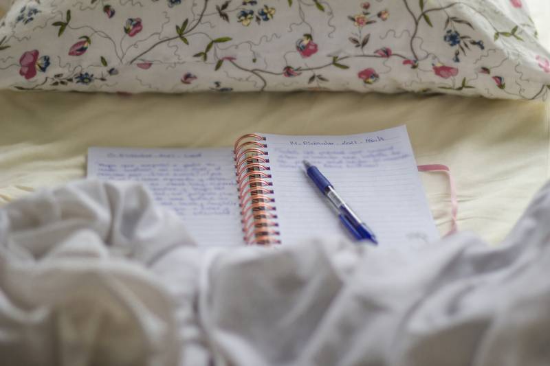 A diary notebook the text out of focus is placed between the sheets of a bed. On top of the pages is a blue pen and on top a flower cushion