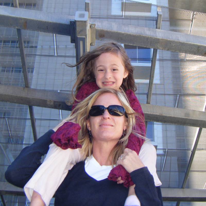 A woman wearing sunglasses holding a small girl on her shoulders outdoors.