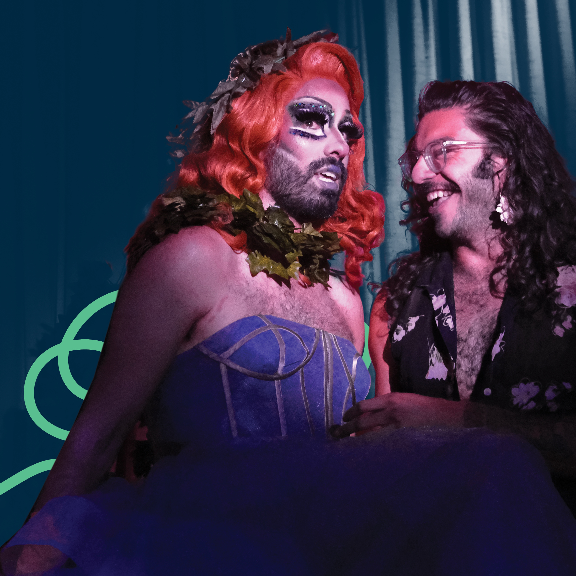 A drag queen with red hair, make-up, and denim top standing next to a smiling, bearded man with glasses and a flower shirt