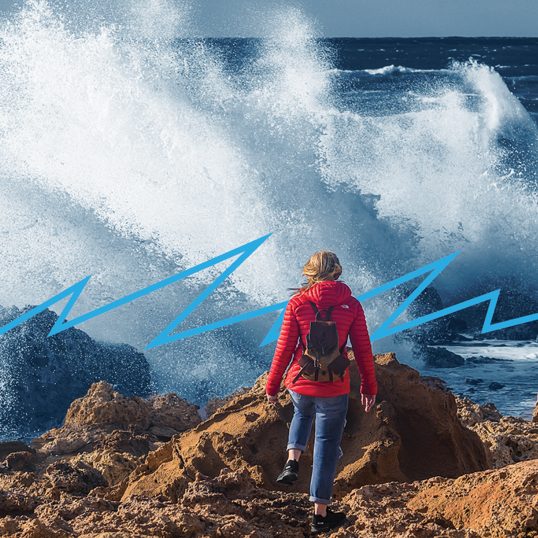 Woman in a red jacket hikes a rocky beach as waves crash in the background