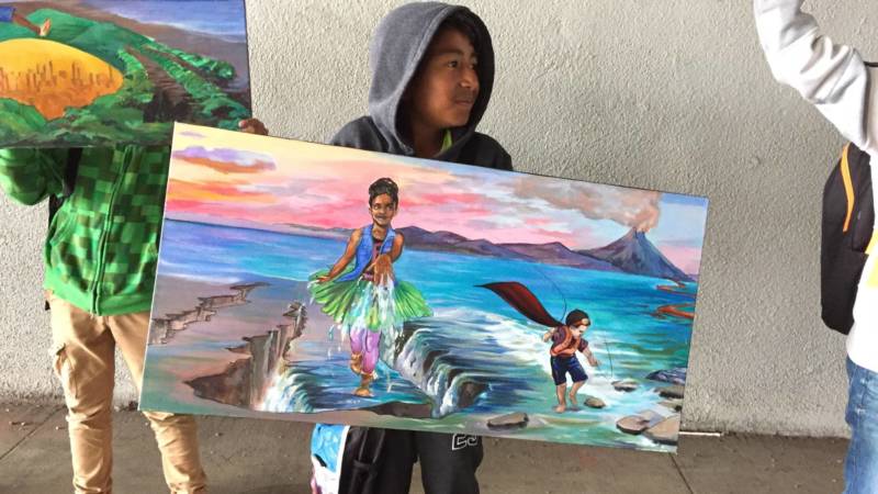 A Hoover Elementary 4th grader holds a canvas showing a section of the planned mural for the I-580 underpass wall behind him