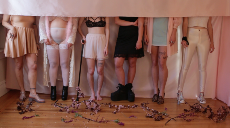 Image description: photo. six people stand facing forward, visible only from the neck down.. a pink fabric hangs and covers their faces in a horizontal line. some use canes. they stand on a wood floor covered with flower petals