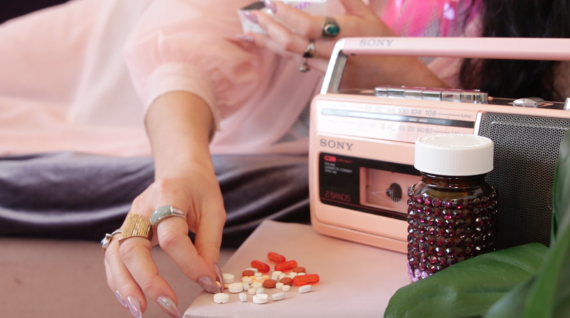 Image Description: photo. a person wearing a light pink nightgown sorts orange and white pills. she has pink opalescent nails. there is a pink radio, a bedazzled pill jar, and a green plant to the right.