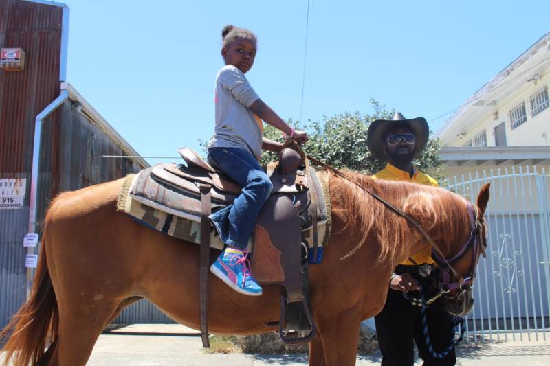 A young lady rides a horse, as a member of the Black Cowboys chaperones her, at a Juneteenth festival in East Oakland.