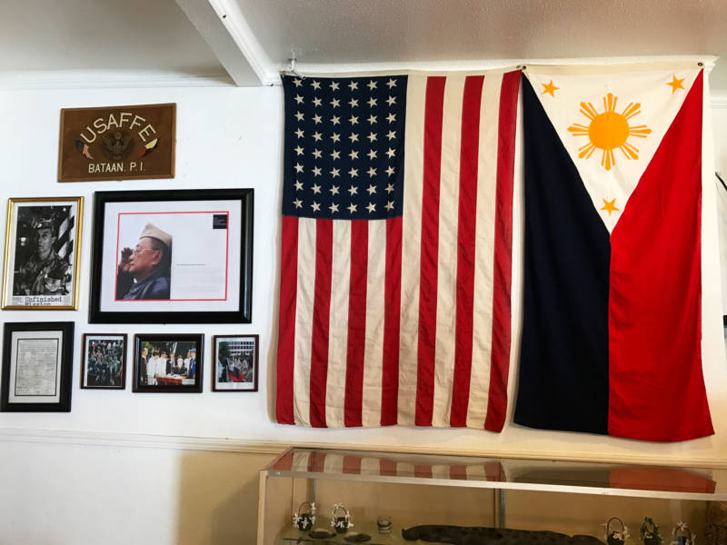 Tess and Marco visit a community center where the flags of the United States and the Philippines are displayed next to images of Filipino veterans of the United States Army Forces in the Far East (USAFFE).
