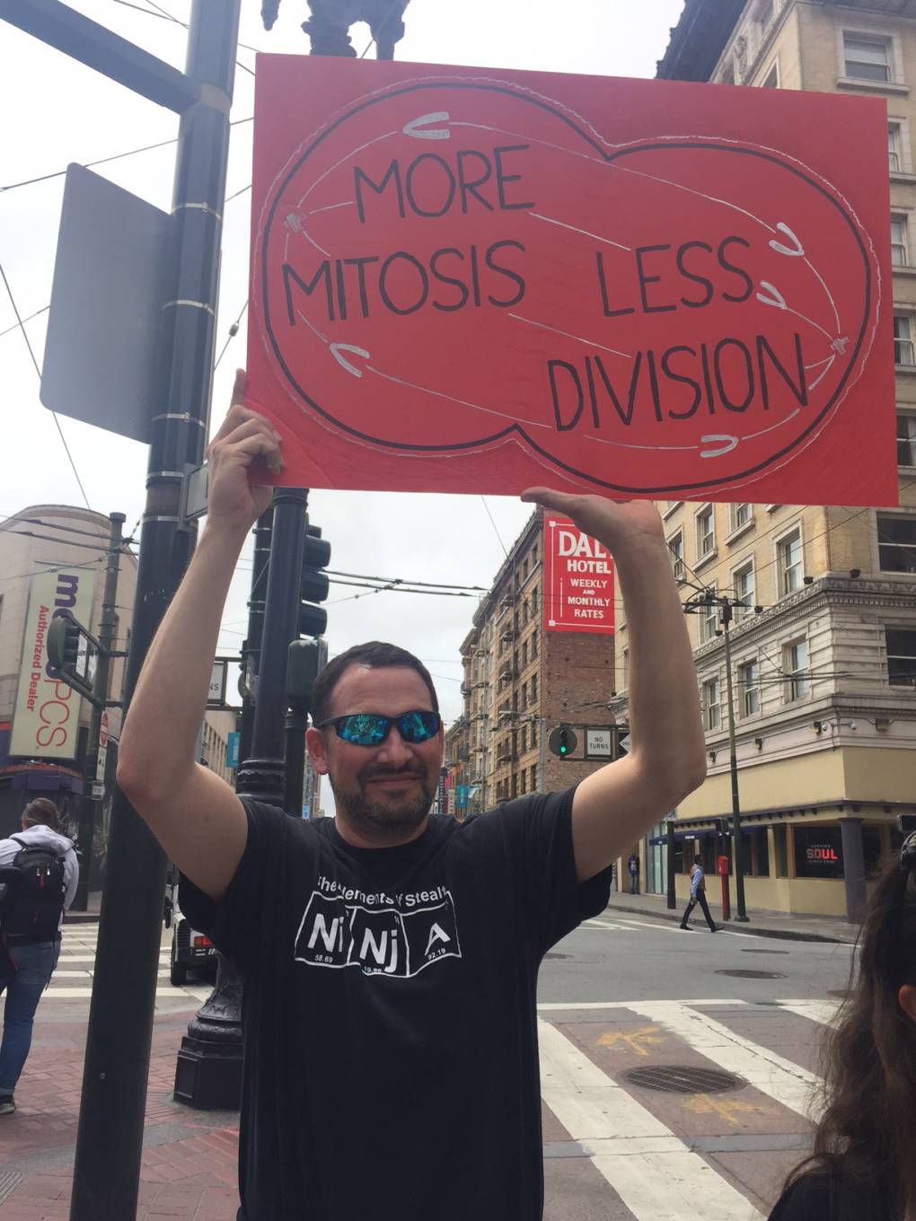 A man holds a sign in Downtown San Francisco that says "More mitosis less division."