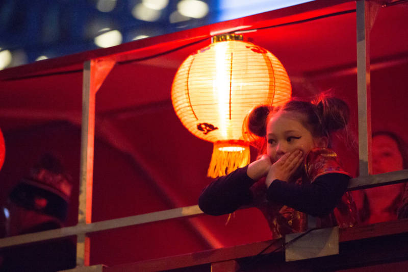 A young girl observes the crowd below on a float decorated with illuminated red and gold lanterns. In traditional Chinese culture, red and gold signify luck and prosperity for the upcoming year.