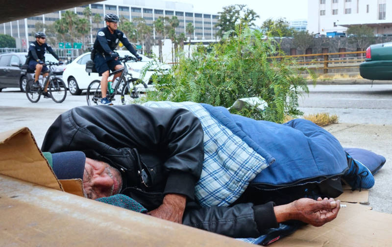 LAPD officers patrol on bicycles past a homeless man in downtown Los Angeles.