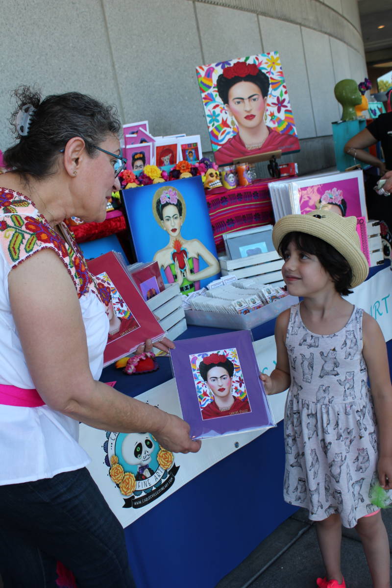 Martha Villanueva of San Jose presents her husband Carlos Villez's artwork to customer Ellie Rue (5) of Walnut Creek at a Frida Kahlo birthday extravaganza in the city on Sunday. Rue says she likes the piece because of "the colors around her hair."