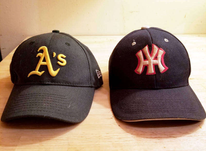 An Oakland A's cap sits next to a Yankee Hater's cap (yes, they exist) on the author's kitchen table.