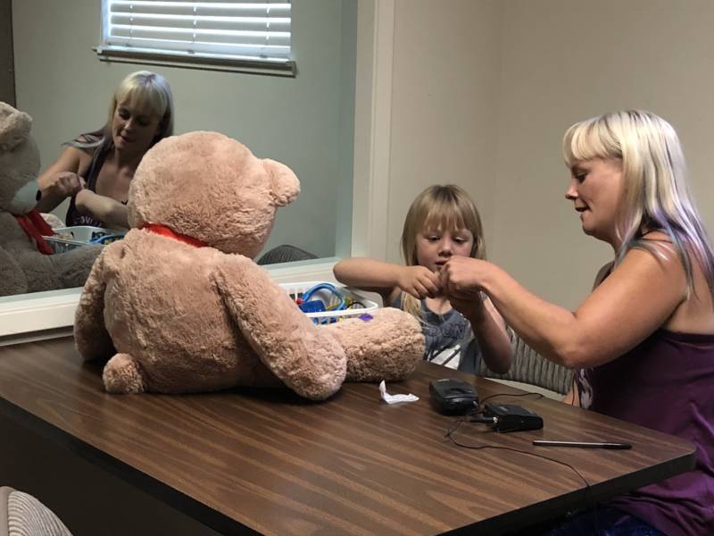 Sabrina Hanes and daughter Aroara during a Parent-Child Interaction Therapy session. Mother and daughter sit on one side of a two-way mirror. A therapist, sitting on the other side of the mirror, coaches Hanes through an earbud she wears.