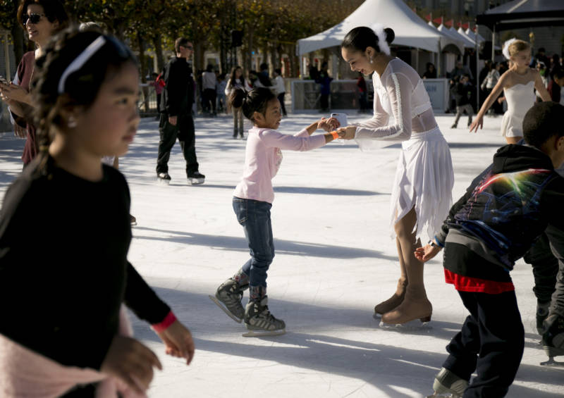 A performer from San Francisco Ice Theatre helps a young girl balance on her skates as other children skate around the rink.