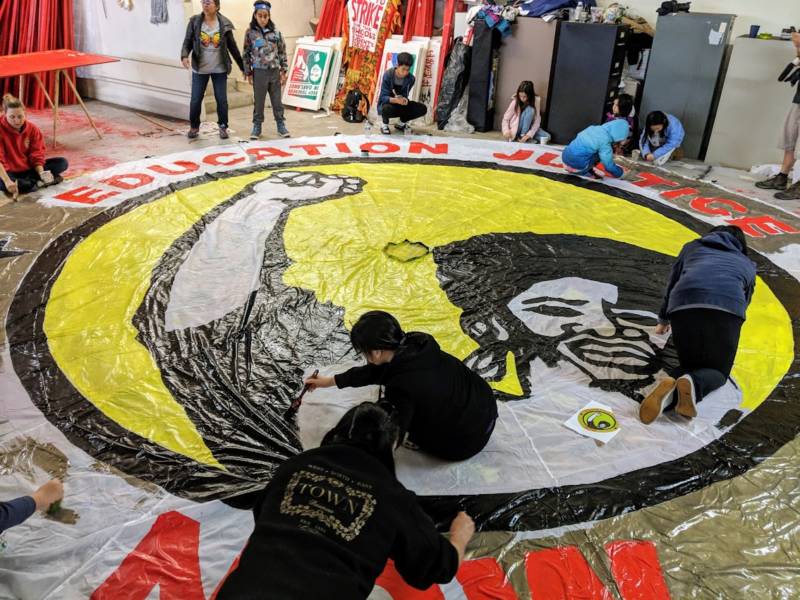 People help paint a parachute that says 'education justice now.' The image is inspired by artwork from Black Panther graphic artist Emory Douglas.