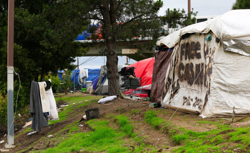 The camp's tents flank a wide transport corridor, situated near BART and Amtrak lines.