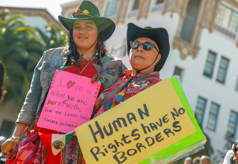 Partners Victoria Miycue and DC Alcalá wear black cowboy hats, hold a small Mexican flag and a yellow sign that say "Human Rights Have No Borders."