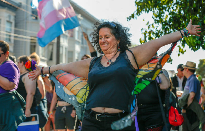 Naomi Tucker, a woman with black curly hair is wearing a black tank top and rainbow-colored butterfly wings.