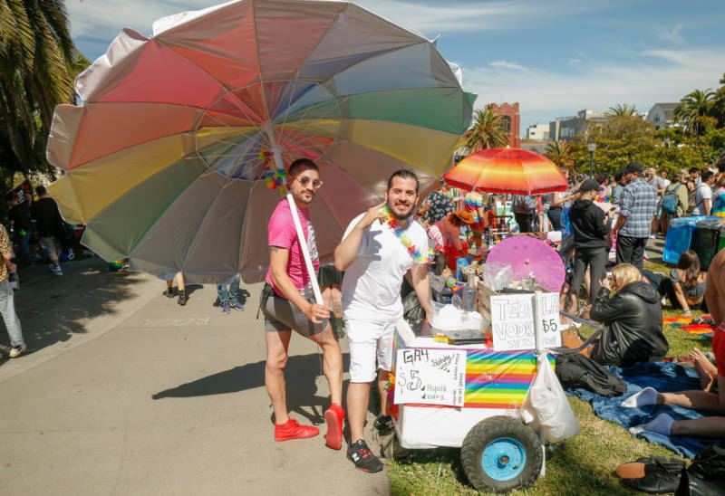 A man waves a huge rainbow umbrella next to an ice cream cart. 