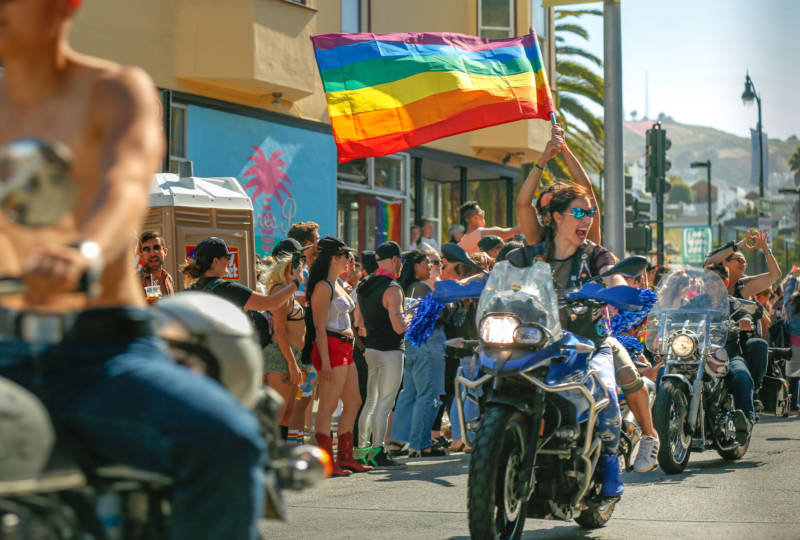 A person on a motorcycle waves a rainbow flag. 