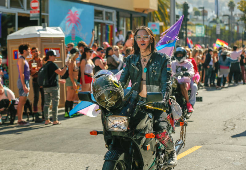 A woman with long hair, wearing a black leather jacket and a bikini top rides her motorcycle down the march. 