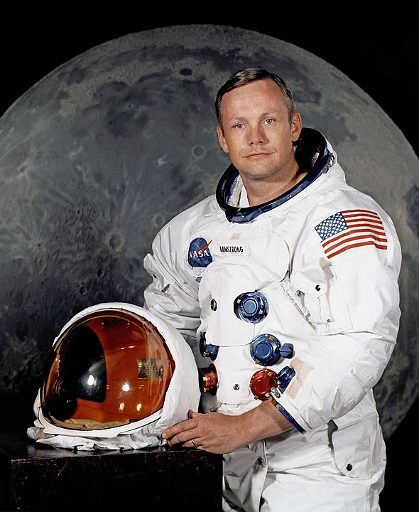 Portrait of Astronaut Neil A. Armstrong, commander of the Apollo 11 Lunar Landing mission in his space suit, with his helmet on the table in front of him. Behind him is a large photograph of the lunar surface.