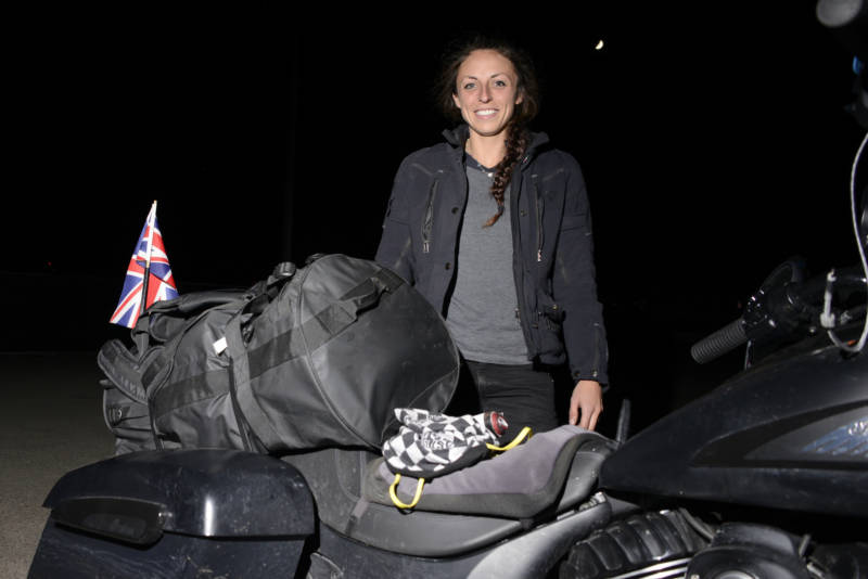 Hayley Bell stands in front of her motorcycle, wearing a black denim jacket.