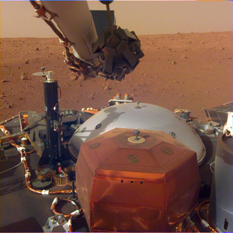 Image of Insight's top deck taken from the Instrument Deployment Camera on the lander's robotic arm. The copper colored object in the foreground is SEIS, Insight's seismometer, which will be placed on the ground to listen for Marsquakes. The gray dome behind SEIS is the wind shield that will be placed on top of it. The black cylinder on the left is HP3, the ground-boring thermal probe that will measure heat flow from within Mars.